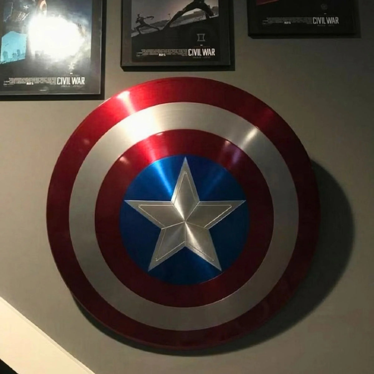 A metallic Captain America shield wall decor piece mounted on a wall, featuring red, white, and blue concentric circles with a silver star in the center. Framed Civil War movie posters are partially visible in the background.